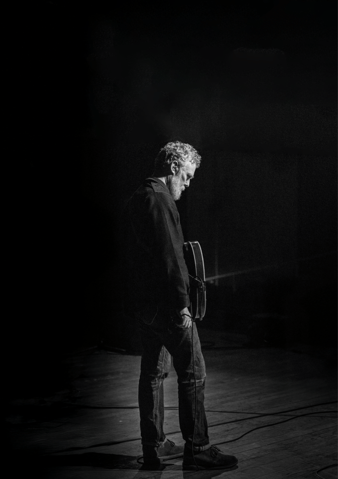Black and white image of Glen Hansard holding a guitar, wearing all black, looking downwards at the ground.